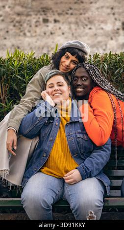 Ripresa verticale di tre diversi giovani amici che sorridono e si abbracciano seduti su una panchina del parco guardando la telecamera. Foto Stock