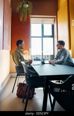 Due uomini sono seduti a una scrivania con laptop in un ambiente d'ufficio moderno e impegnati in una conversazione. La camera presenta un decor minimalista e un design naturale Foto Stock