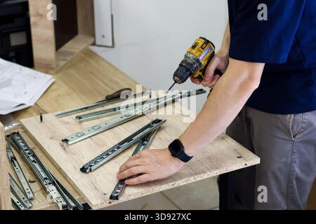 Persona che fissa le guide metalliche del cassetto al pannello di legno utilizzando un trapano a batteria. Operaio in camicia blu che fora viti nelle guide per cassetti su furnitu in compensato Foto Stock