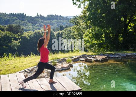 Donna attiva e matura durante la routine mattutina yoga posizione guerriera presso la piscina naturale in giardino. Relax esercizio all'aperto e stile di vita sano Foto Stock