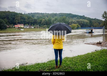 Donna con ombrello guarda il fiume durante l'alluvione. Calamità naturali e danni ambientali dopo piogge torrenziali Foto Stock