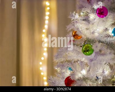 Primo piano di un albero di Natale artificiale bianco con ornamenti vivaci e luci festive. Temi festivi, invernali e festeggiamenti. Foto Stock