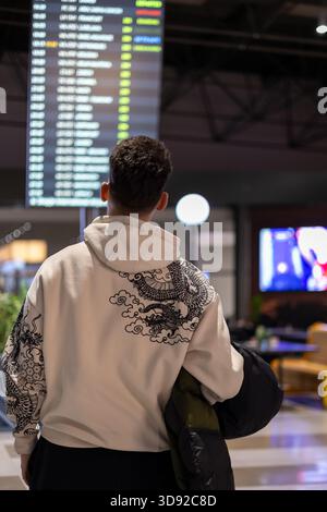 un giovane con una felpa con cappuccio bianca si erge con le spalle alla telecamera, guardando un luminoso bordo di partenza in un moderno terminal aeroportuale Foto Stock
