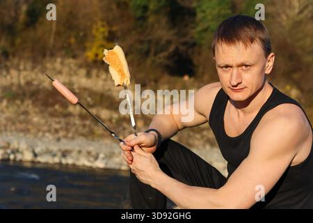 Uomo determinato in camicia senza maniche che tiene saldamente spiedini di hot dog grigliato in riva al fiume. Foto Stock