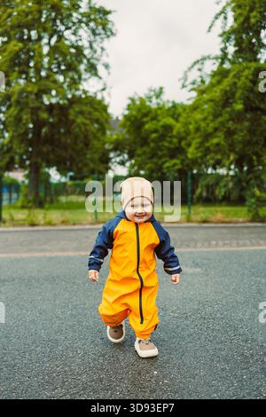 Un vivace bambino vestito da accattivante impermeabile giallo cammina con gioia lungo un sentiero lastricato in un parco circondato da vegetazione lussureggiante e cieli ricoperti, Foto Stock