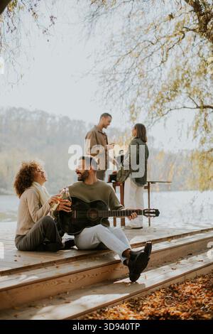 Quattro amici si riuniscono vicino all'acqua in una giornata calda, gustando bevande, risate e musica mentre si rilassano sul ponte di legno. Foto Stock