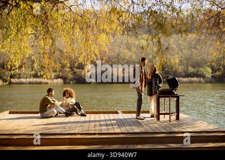 Quattro amici si riuniscono su una piattaforma di legno che si affaccia su un fiume calmo, condividendo risate e grigliando cibo delizioso sotto foglie vivaci. Foto Stock