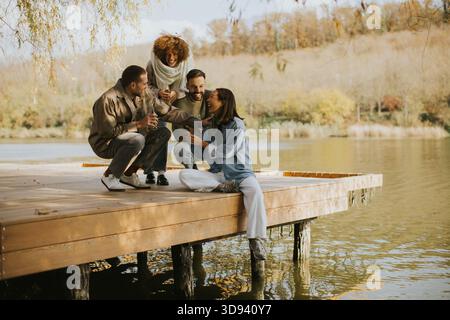 Un gruppo di quattro amici si riunisce su una banchina di legno, condividendo bevande e risate circondati da foglie autunnali e acque calme. Foto Stock