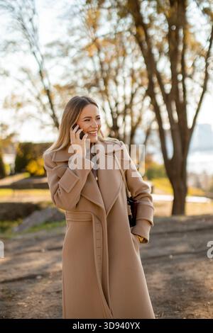 Una giovane donna allegra con un cappotto elegante sta conversando sul suo telefono. Si trova in un parco circondato da alberi con foglie autunnali. La luce del sole aggiunge wa Foto Stock