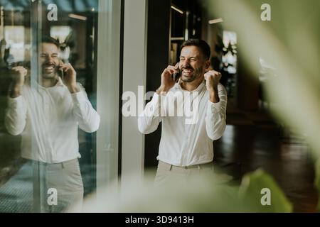 Un uomo che indossa una camicia bianca sorride molto e alza i pugni di gioia mentre parla al telefono. Si trova all'interno di un ufficio moderno con grandi finestre Foto Stock