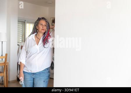 Donna di mezza età con dreadlock colorati appoggiati su un muro bianco nella sua casa illuminata, che guarda con attenzione lontano. Foto Stock