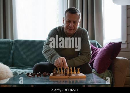 Uomo anziano concentrato seduto a casa a giocare a scacchi, godendo di una tranquilla attività al coperto e di un piacevole tempo libero. Home attività ricreative Foto Stock