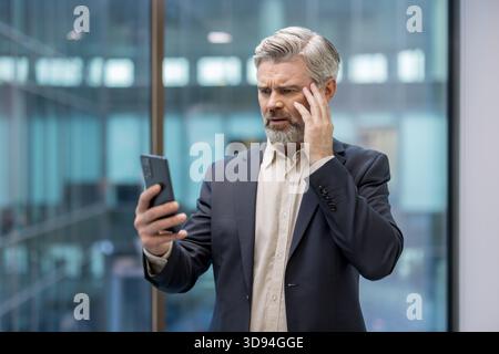 Uomo d'affari anziano che riceve brutte notizie inaspettate sul suo cellulare, mostrando un'espressione preoccupata e confusa con la mano alla testa in un moderno ufficio Foto Stock