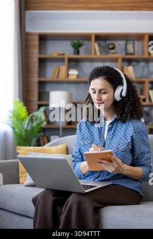 Giovane donna su un divano che indossa le cuffie e lavora su un notebook, prendendo appunti scritti a mano in un notebook mentre frequenta una lezione online dal suo salotto di casa Foto Stock