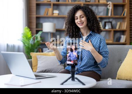 Donna che crea contenuti online, parla con la fotocamera durante un live streaming o una videochiamata di successo da casa, utilizzando uno smartphone su un cavalletto con un laptop presente Foto Stock