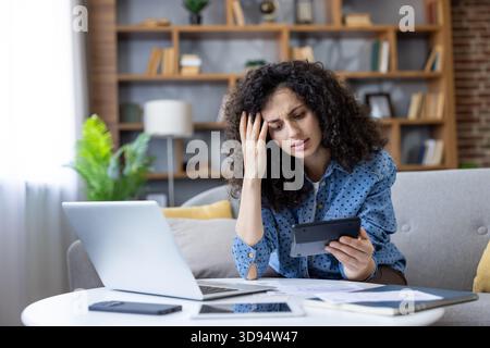 La donna si sente stressata e preoccupata durante il calcolo delle spese familiari, delle bollette e dell'analisi di documenti e fatture con una calcolatrice e un notebook a casa, con difficoltà finanziarie Foto Stock