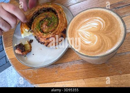 Primo piano di mani che condividono una sfoglia di cannella e pistacchio con una grande tazza di caffè latte art Foto Stock