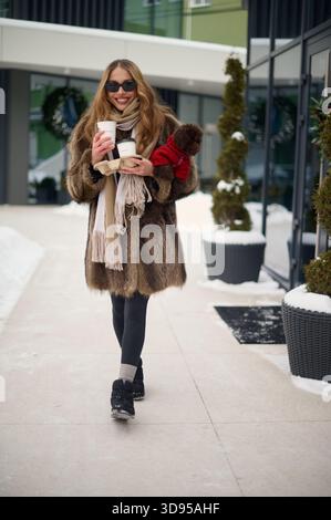 Donna felice e alla moda con occhiali da sole e un cappotto di pelliccia con due tazze di caffè da asporto e un piccolo cane marrone vestito con un maglione rosso mentre sei in piedi Foto Stock