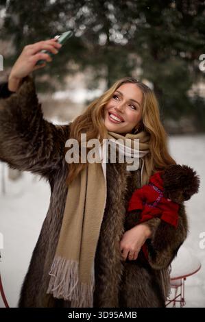 Una donna gioiosa, vestita con un comodo cappotto di pelliccia e sciarpa, scatta un selfie all'aperto in un ambiente innevato, tenendo un piccolo cane in un maglione rosso. Foto Stock