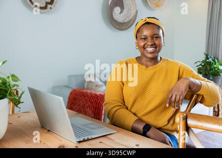 Donna afroamericana che indossa un maglione giallo che scrive su un computer portatile a un tavolo vicino alle piante in salotto Foto Stock