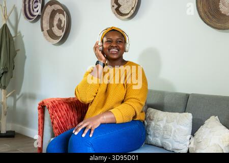 Donna afroamericana seduta sul divano di casa con maglione giallo e fascia con cuffie Foto Stock