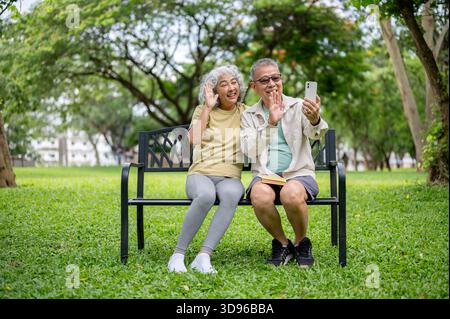 Occhiali vecchio che tiene il telefono a fare videochiamate a parte donna sorridente mentre si siede insieme sulla panchina nel parco. Coppia di anziani, natura, relax all'aperto. Foto Stock