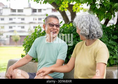Un vecchio di vetro sorride e parla con una vecchia mentre si siede insieme sulla panchina di cemento nel parco. Coppia di anziani, Green Nature, Chilling Outdoor. Foto Stock