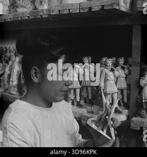 Slavyansk, Ucraina, URSS. 1968. Foto d'archivio in bianco e nero che mostra una dipendente della mietitrebbia Slavyansk Ceramic che esegue il controllo di qualità sui prodotti in porcellana. La donna con un cappotto da lavoro bianco ispeziona da vicino una piccola figurina tra le sue mani. Dietro di lei, gli scaffali di legno sono riforniti di file di statue identiche raffiguranti ragazze in cappotti e muff invernali. L'immagine illustra la produzione di massa di souvenir di consumo e l'attenzione ai dettagli nel pacifico Donbass prima della guerra. Foto Stock