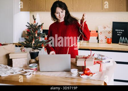 Una giovane donna sorride mentre usa il laptop e tiene il nastro accanto all'albero di Natale decorato e ai regali. Workshop virtuale di Natale, vacanze Foto Stock