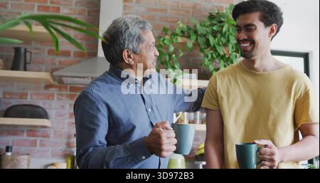In mano tazze, uomo anziano con bottone-down blu e uomo adulto con maglietta gialla che chatta in cucina Foto Stock