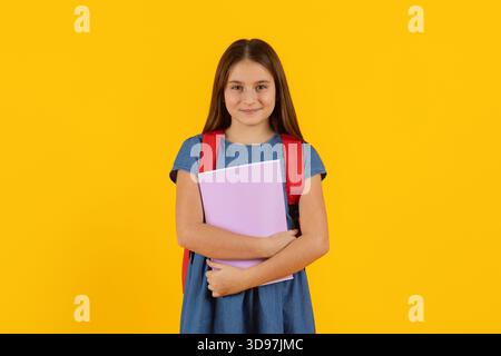 Ragazza preadolescente in piedi con zaino e notebook sullo sfondo giallo brillante dello studio Foto Stock