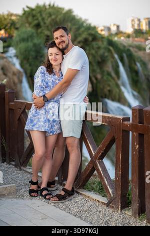 Una coppia si erge a stretto contatto con una ringhiera di legno, sorridente e avvolgente con splendide cascate sullo sfondo, creando un'atmosfera tranquilla Foto Stock