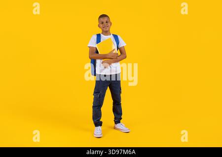 Preadolescente in piedi con notebook e zaino sullo sfondo giallo brillante dello studio Foto Stock