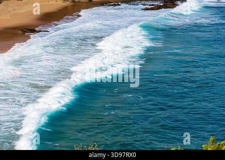 Una potente onda oceanica con una brillante cresta turchese si schianta, creando un'acqua bianca spumeggiante contro una spiaggia di sabbia dorata. Foto Stock