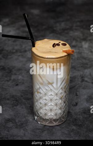 Un alto vetro di cristallo a motivi geometrici contiene un latte a strati ricoperto di schiuma cremosa, accentuato da una cannella e da chicchi di caffè. Ricco, invitante e. Foto Stock