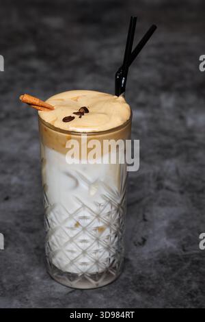 Un alto vetro di cristallo a motivi geometrici contiene un latte a strati ricoperto di schiuma cremosa, accentuato da una cannella e da chicchi di caffè. Ricco, invitante e. Foto Stock