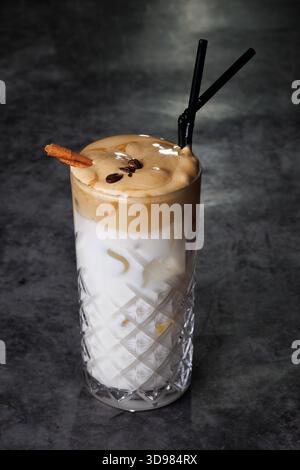 Un alto vetro di cristallo a motivi geometrici contiene un latte a strati ricoperto di schiuma cremosa, accentuato da una cannella e da chicchi di caffè. Ricco, invitante e. Foto Stock