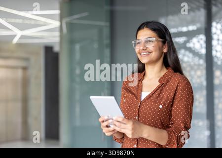 Giovane donna d'affari indiana che utilizza un tablet digitale e sorride con fiducia, in un ambiente d'ufficio moderno, che rappresenta una carriera di successo e. Foto Stock