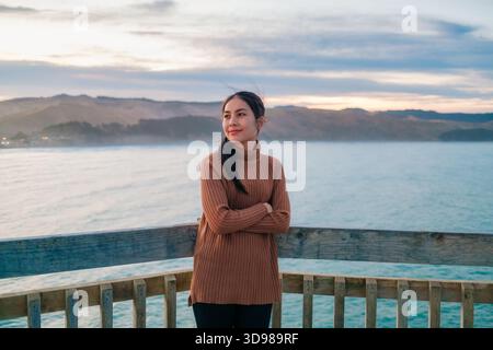 Bella donna asiatica in maglione marrone in piedi sul lungomare del pacifico con vista sulle montagne al tramonto a Wellington, nuova Zelanda Foto Stock