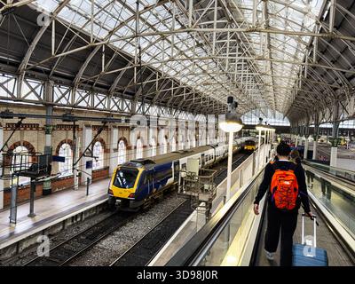 Manchester, Regno Unito - 7 novembre 2025: Un viaggiatore con uno zaino arancione cammina lungo un passaggio pedonale in movimento verso un treno blu alla stazione di Manchester. Foto Stock