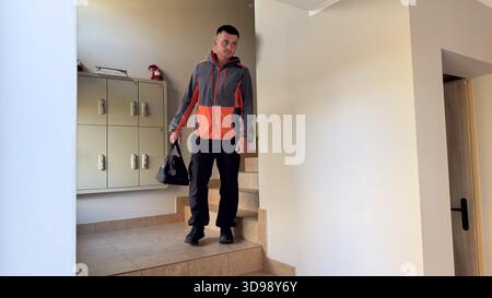Un uomo con una giacca grigia e arancione scende le scale piastrellate in un corridoio dell'appartamento, portando un borsone nero vicino a un armadietto a parete e a un labbro Foto Stock