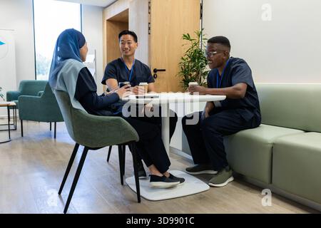 Diversi colleghi medici seduti in sala pausa indossando scrub della marina tenendo tazze di caffè e telefono Foto Stock