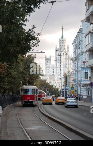 Mosca, Russia - 6 settembre 2025: Un tram rosso si sposta lungo i binari della città con auto ed edifici storici, grattacielo stalinista che si innalza sullo sfondo Foto Stock