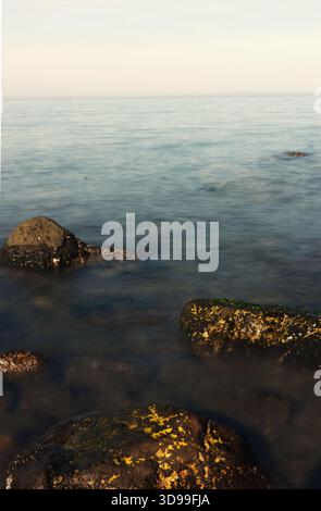 Primo piano verticale di Mossy Rocks su una linea costiera calma e sfocata Una composizione verticale e ravvicinata che si concentra su grandi rocce scure parzialmente sommerse nel mare Foto Stock