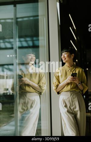Una donna con camicetta verde chiaro e pantaloni beige si erge accanto a una grande finestra in vetro in un moderno caffè. Sorride mentre guarda il suo smartphone, rif Foto Stock