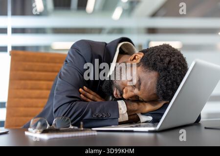 Uomo d'affari afroamericano addormentato alla sua scrivania con laptop e occhiali, esausto e sovraccarico di lavoro, che illustra l'esaurimento del luogo di lavoro, la stanchezza cronica e lo stress in un ufficio aziendale Foto Stock