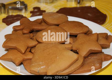 Biscotti a forma di stelle, campane e altre figure sono disposti su un piatto bianco. I biscotti sono visibili sullo sfondo. Questa scena ha luogo Foto Stock