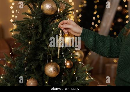Primo piano di ornamenti dorati appesi a mano sull'albero di Natale con luci calde in un intimo interno natalizio. Foto Stock