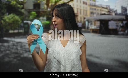 Giovane donna bruna che tiene un grande nastro blu di consapevolezza nel mercato di strada acciottolato, bocca aperta con gesto sorpreso; sostenere la consapevolezza sorpreso. Foto Stock