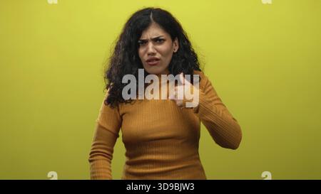 Giovane donna bruna che punta i pollici sul petto indossando un maglione senape in studio giallo; dubbio di sé confusione. Foto Stock